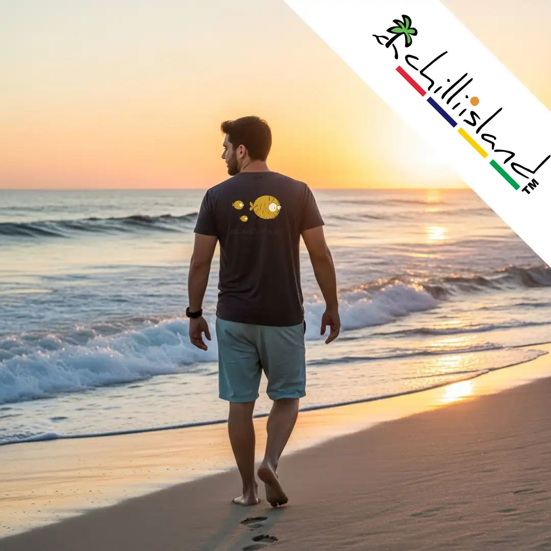 Man walking on a beach at sunrise, wearing a dark shirt with a Chilliisland Island Punk design. Waves gently crash, creating a serene and peaceful atmosphere.