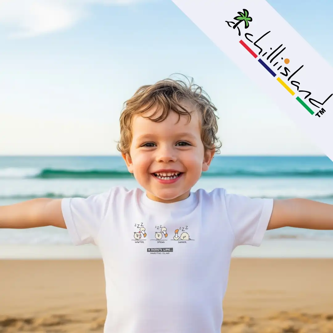 A child stands with arms outstretched on a beach, wearing a white t-shirt featuring a playful cartoon design Dodo Life. Ocean waves are in the background.